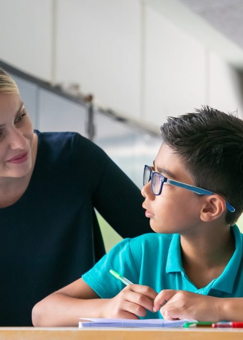 Cheerful school teacher giving help and support to schoolboy in class. Front view. Education or back to school concept